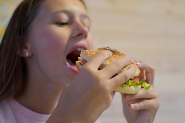 食欲のある女の子は美味しいハンバーガーを食べます。子供はサンドイッチの大きな部分をかみます