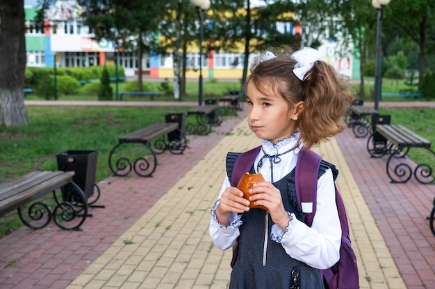 バックパックを背負って学校の近くでパイを食べる女の子 学校からのパンで簡単なおやつ 不健康な食べ物 学校に戻る 教育 小学校の授業 9 月 1 日
