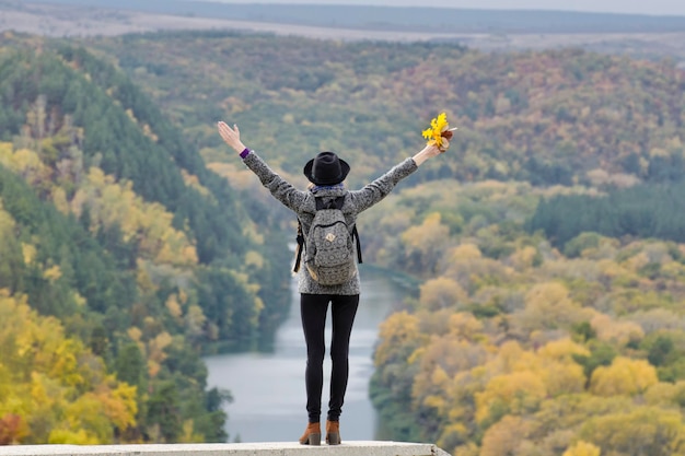 Girl with a backpack and a hat standing on a hill Hands raised up River and mountains below Back view