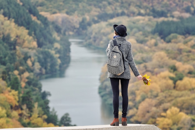 Girl with a backpack and a hat standing on a hill. River and forest below. Back view