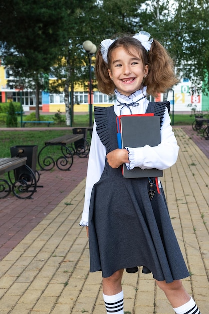Girl with backpack school uniform with white bows and stack of books near school Back to school happy pupil heavy textbooks Education primary school classes September 1