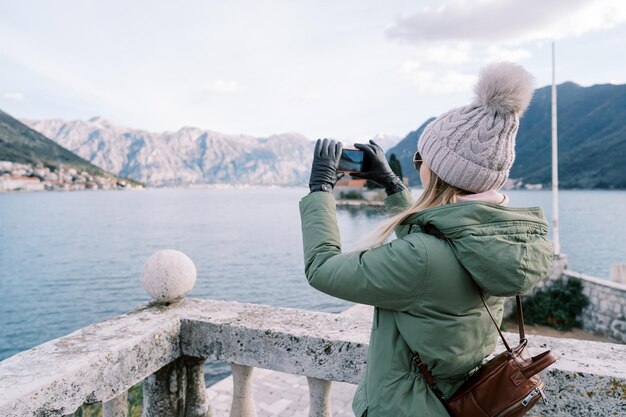 Photo girl with a backpack shoots the sea and a mountain range with a smartphone while standing at a stone