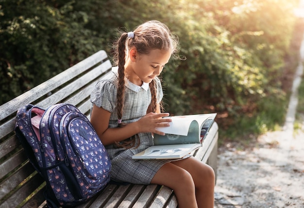 Girl with a backpack sitting on a bench and reading a book near\
the school back to school lesson schedule a diary with grades\
education primary school classes back to school