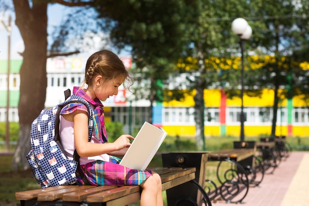 Girl with a backpack sitting on a bench and reading a book near the school. Back to school, lesson schedule, a diary with grades. Education, primary school classes, September 1