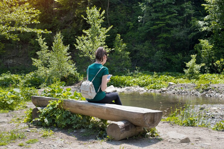 Girl with a backpack and a smartphone sitting on a river bank. Summer sunny day