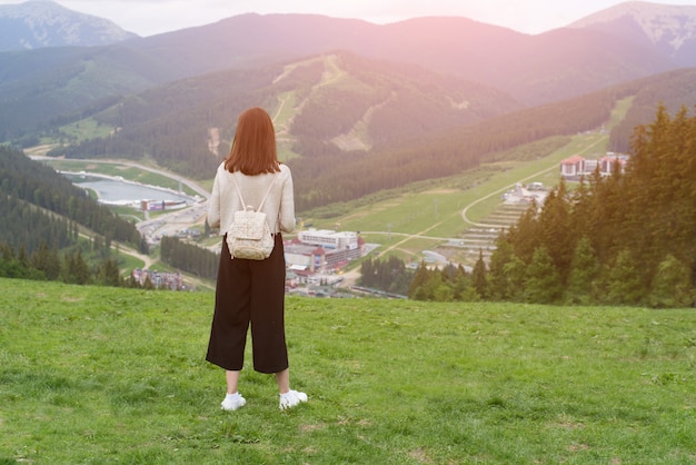 丘の上に立って、山を眺めるバックパックを持つ少女。遠くの町