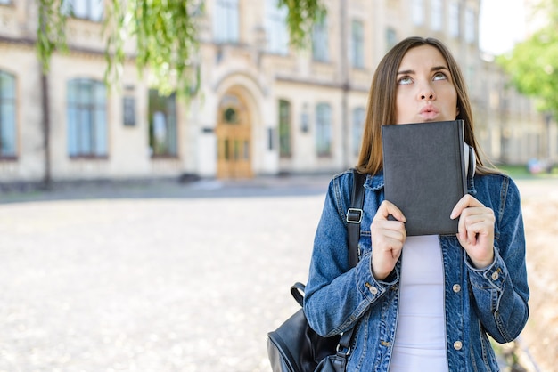 Девушка с сумкой и книгой смотрит на фоне кампуса