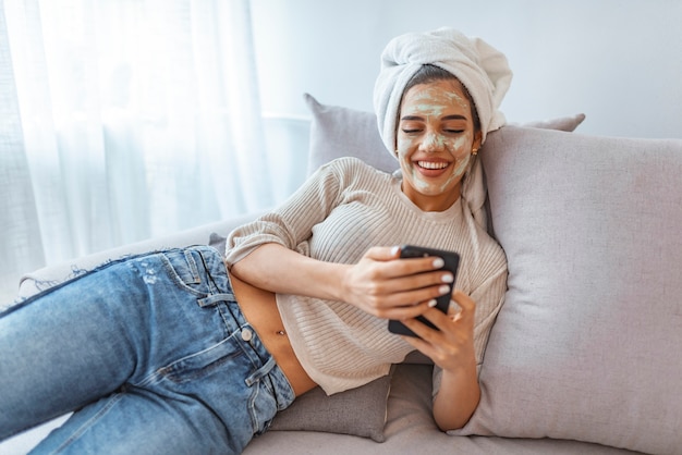 Foto ragazza con la maschera di bellezza che si rilassa a casa, testo di invio di messaggi sul telefono cellulare