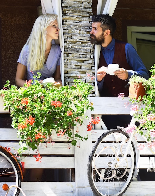 Girl with blond hair and bearded guy on cafe background. Morning date concept. Woman and man with dreamy faces have a date on terrace with flowers and bike. Couple in love holds cups of coffee outside