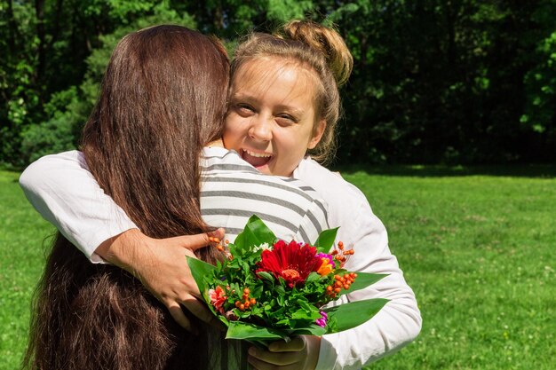 A girl with blond hair combed in a bun gives her mother a bouquet of red flowers