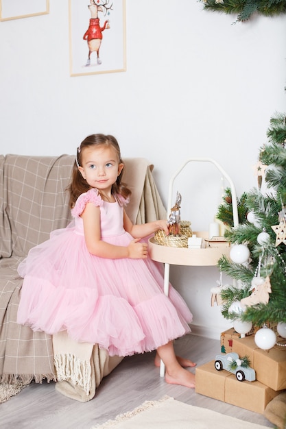 Girl with blond hair in a smart pink dress near the Christmas tree sits on the sofa and plays