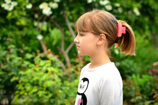 A girl with blond hair stands on a background of green leaves.