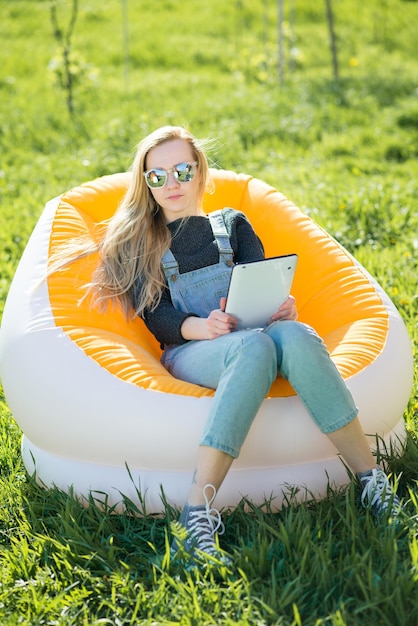 Girl with blond long hair with a tablet sitting on an inflatable chair on a green lawn