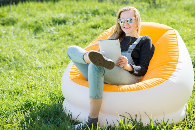 Девушка со светлыми длинными волосами с планшетом сидит на надувном стуле на зеленой лужайке