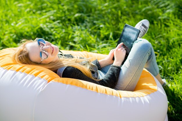 Photo girl with blond long hair with a tablet sitting on an inflatable chair on a green lawn