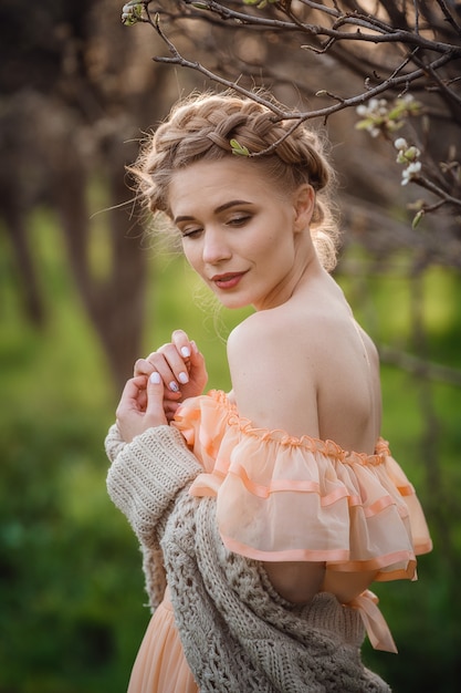 Girl with blonde hair in a light dress in flowering garden. concept of female spring fashion. girl in a beautiful dress and knitted sweater enjoys the sunset in a pear flowering garden