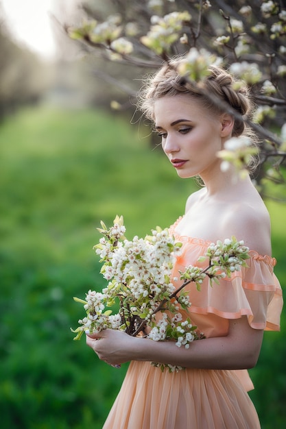 花の咲く庭で明るいドレスを着たブロンドの髪の少女。女性の春のファッションのコンセプト。