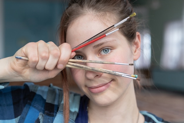 A girl with blue eyes and red hair holds in her hand many brushes and looks through them. Woman in the process of drawing oil paintings.