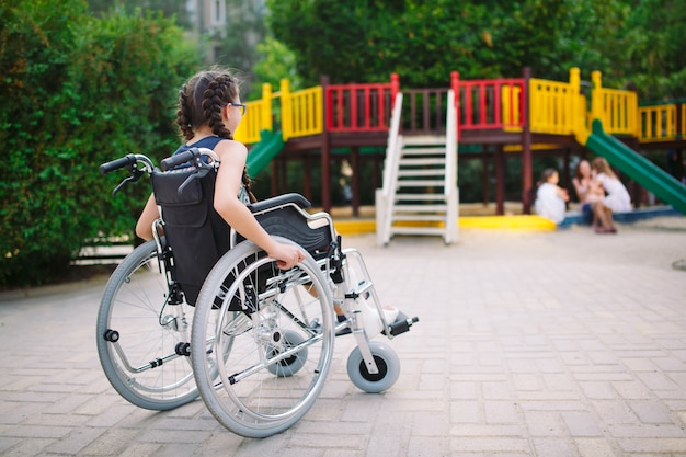 Девушка со сломанной ногой сидит в инвалидной коляске перед детской площадкой.