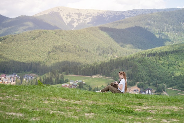 Girl with the camera sits on a hill and viewing photos. Mountains in the background