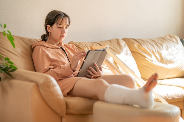 다리에 깁스를 하고 소파에 앉아 그래픽 태블릿으로 그림을 그리는 소녀 달리기 부상 사고 후 붕대를 감은 다리 깁스와 발가락 석고 깁스를 한 십대 소녀