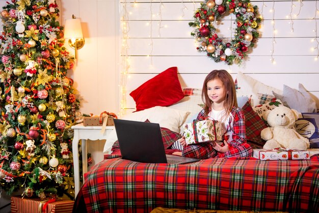 A girl with a Christmas gift in her hands sits in her pajamas on a bed opposite a laptop in a decorated bedroom.