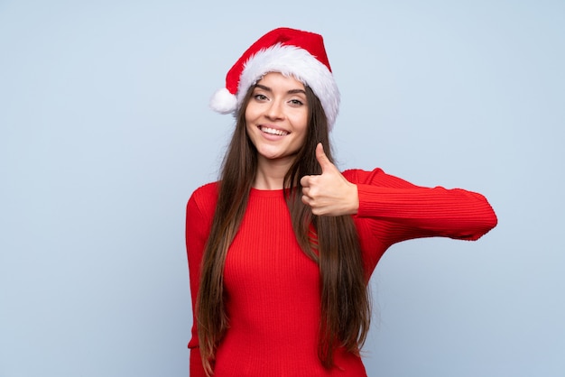 Girl with christmas hat over isolated blue giving a thumbs up gesture