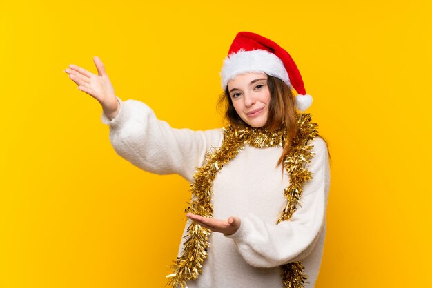 Girl with christmas hat over isolated yellow wall extending hands to the side for inviting to come