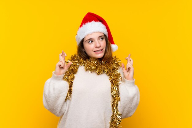 Girl with christmas hat over isolated yellow wall with fingers crossing