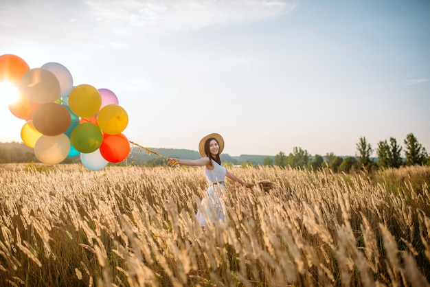 Ragazza con palloncini colorati che camminano nel campo di grano. bella donna sul prato estivo al giorno pieno di sole