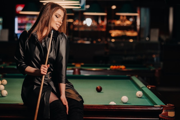 A girl with a cue in her hands sits on a table in a billiard club.Russian billiards