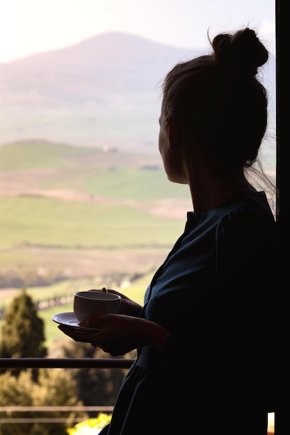 커피 한 잔을 들고 있는 소녀가 창 밖 창가에 서 있습니다. 토스카나 풍경입니다. 투스카니, 이탈리아