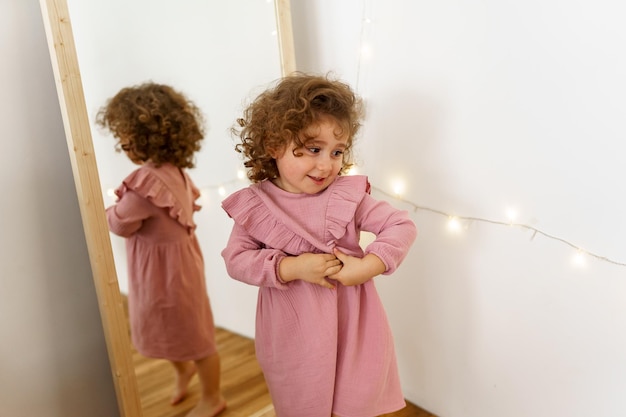 a girl with curls in a pink dress laughs at the mirror