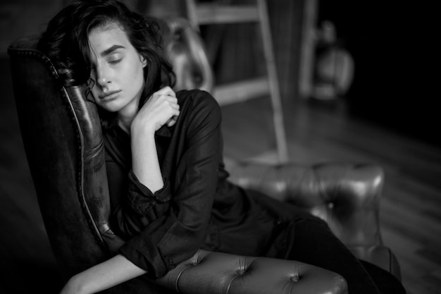 Girl with curly dark hair and natural makeup sitting on a dark leather chair black and white photo