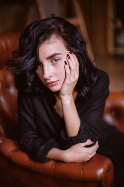 Girl with curly dark hair and natural makeup sitting on a leather chair vintage photo processing
