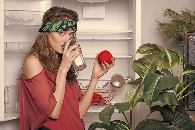 Girl with curly hair drinking milk Woman wearing headband holding red apple vitamins and health concept Lady in oversized silk blouse standing next to open fridge Big green plant in white kitchen