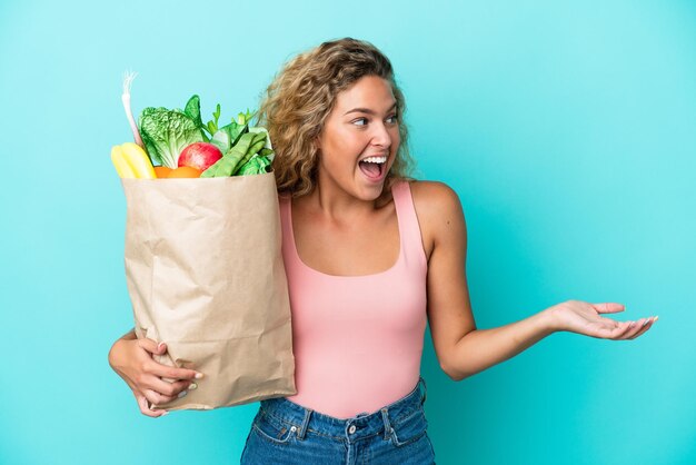 Girl with curly hair holding a grocery shopping bag isolated on green background with surprise expression while looking side