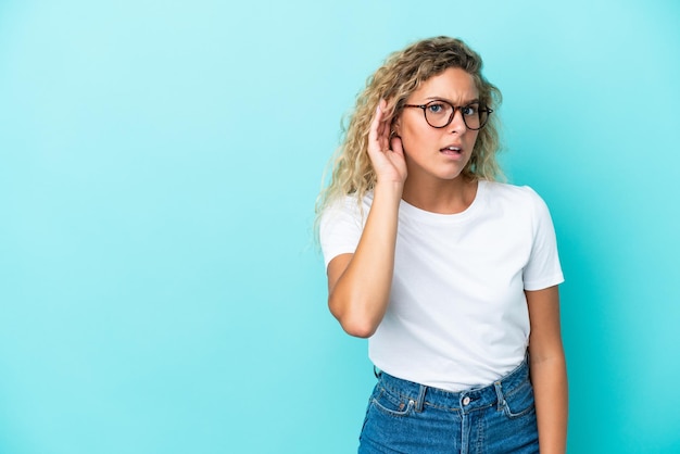 Girl with curly hair isolated on blue background listening to something by putting hand on the ear