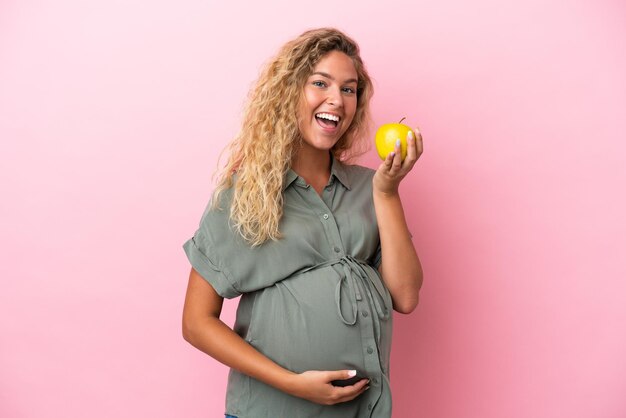 Девушка с вьющимися волосами на розовом фоне беременна, держит яблоко и ест его