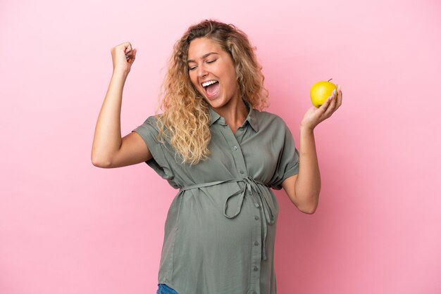 Girl with curly hair isolated on pink background pregnant and holding an apple while celebrate a victory