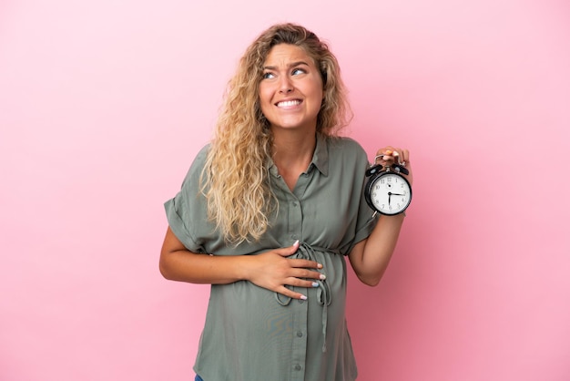 妊娠中のピンクの背景に分離され、ストレスの多い表現で時計を保持している巻き毛の女の子