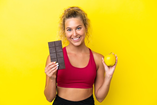 Girl with curly hair isolated on yellow background taking a chocolate tablet in one hand and an apple in the other