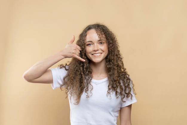 A girl with curly hair shows a call me gesture on a beige background