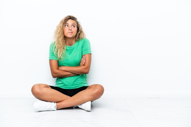 Foto ragazza con i capelli ricci seduta sul pavimento che fa un gesto di dubbi mentre si sollevano le spalle