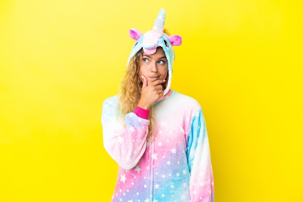 Girl with curly hair wearing a unicorn pajama isolated on yellow background having doubts and with confuse face expression