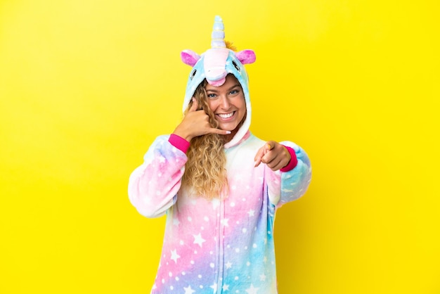 Girl with curly hair wearing a unicorn pajama isolated on yellow background making phone gesture and pointing front