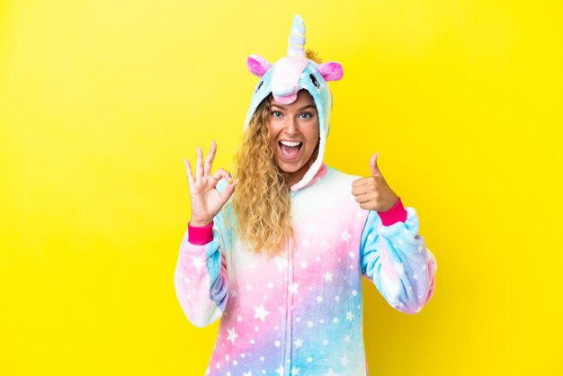 Girl with curly hair wearing a unicorn pajama isolated on yellow background showing ok sign and thumb up gesture