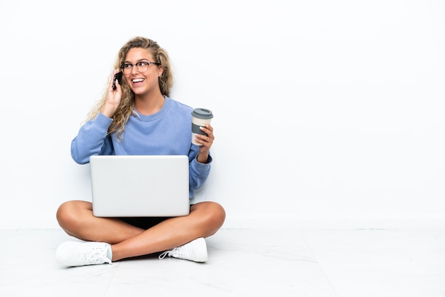 Girl with curly hair with a laptop sitting on the floor holding coffee to take away and a mobile