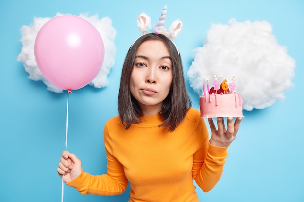 La ragazza con i capelli scuri tiene in mano una deliziosa torta di fragole e il palloncino gonfiato sembra calmo alla telecamera indossa abiti casual isolati su blu