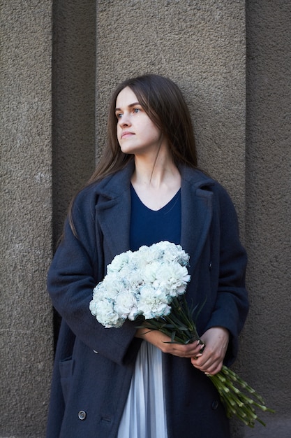 A girl with dark hair holds in her hands a bouquet of blue carnations on gray wall 